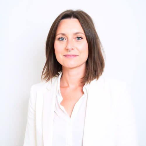 Frau mit blonden Haaren und Piercing in einem weißen Blazer vor weißem Hintergrund.