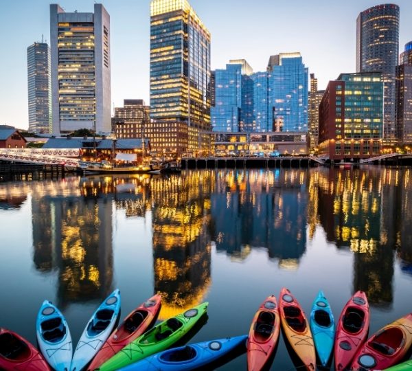 Boston Skyline am Hafen im Abendlicht spiegelt sich im ruhigen Wasser; bunte Kajaks im Vordergrund prägen das Bild mit moderner Stadtsilhouette