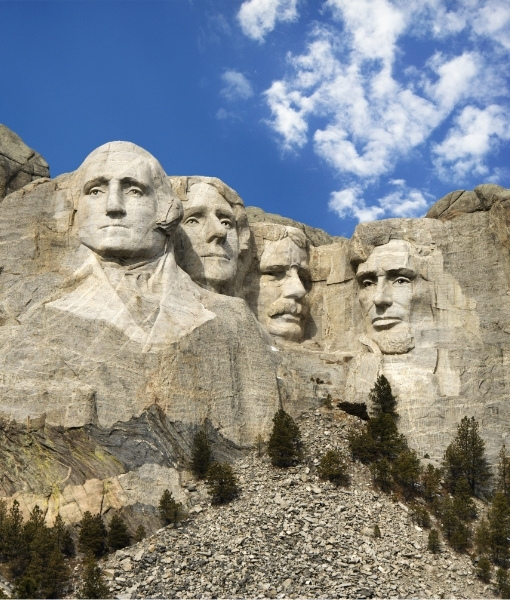 Monumentale Mount-Rushmore-Figuren aus hellgrauem Stein vor blauem Himmel mit Wolken, eingebettet in eine felsige Landschaft des Mittleren Westens.
