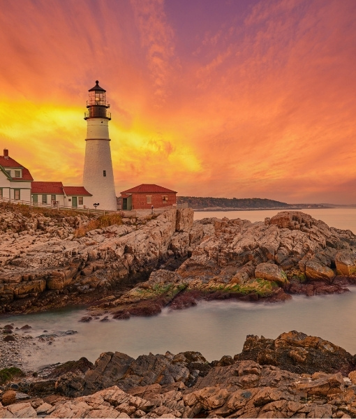 Leuchtturm an felsiger Küste bei Sonnenuntergang an der Ostküste USA, umgeben von Wasser und Steinen, mit orangefarbenem Himmel und dramatischem Licht.