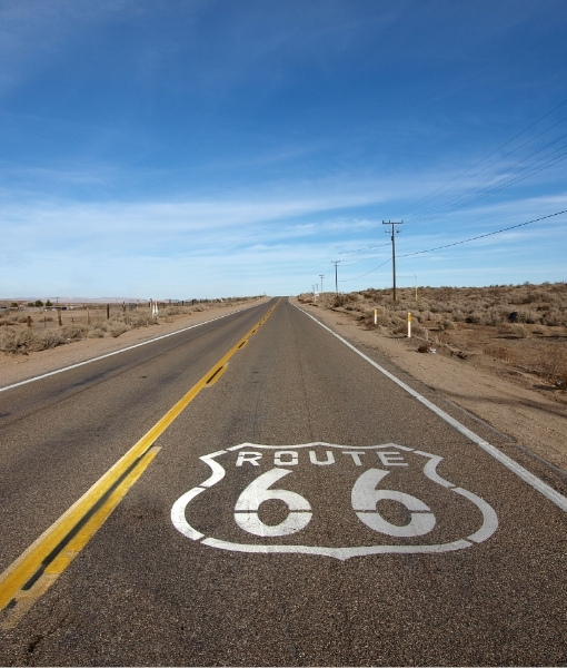 Weite Straße mit gelber Mittelmarkierung und weißer ROUTE-66-Beschriftung durch die trockene Landschaft, begleitet von Stromleitungen am Straßenrand.