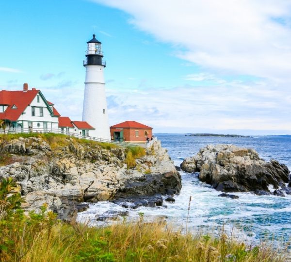 Portland, Maine – weißer Leuchtturm am felsigen Küstenstreifen, rotes Backsteinhaus, graue Felsen und blauer Himmel, Atlantik-Inselidyll