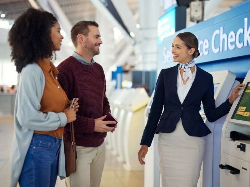 Lächelnde Reisende nutzen den Online-Check-in am Terminal, während eine Mitarbeiterin am Automaten hilft, mit blauem Bildschirm im Hintergrund.