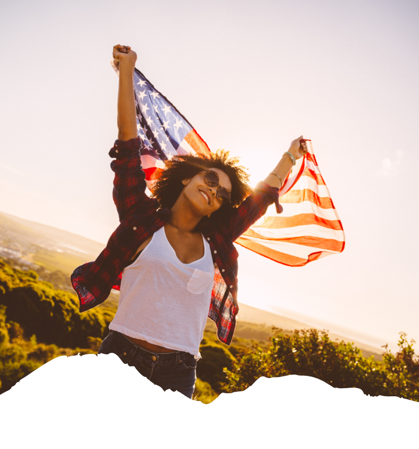 Sonniges Outdoor-Motiv mit US-Flaggenmuster im Wind: Eine Person hebt die Arme auf einer Anhöhe und zeigt die USA-Flagge vor weitem Panorama.