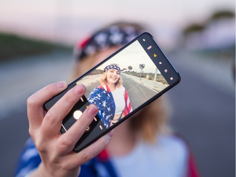 Hand hält ein Smartphone mit Ansicht einer Person vor einer amerikanischen Straße und Flaggenmotiv in blau und rot-weiß, passend zum eSim-USA Angebot.