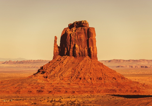In der warmen Abendsonne ragt eine markante rote Felsformation über die weite Wüstenebene Arizonas auf, eingerahmt von Himmel und Fernblick.