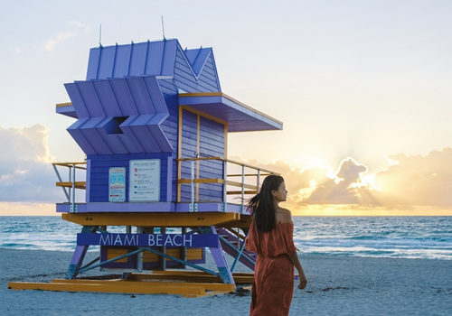 Am Miami Beach steht ein lilafarbener Rettungsturm vor dem Meer, während eine Frau im roten Kleid am Strand entlanggeht und die Sonne untergeht.