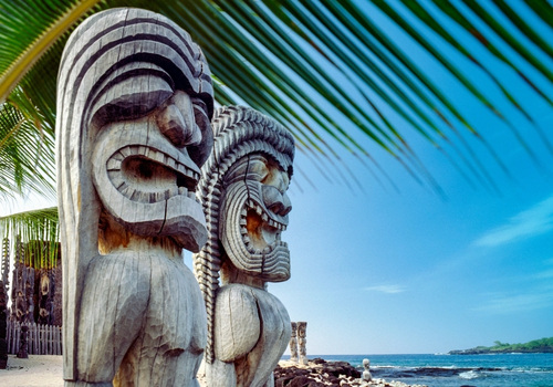 Vor blauem Himmel stehen zwei geschnitzte Tiki-Statuen an einer hawaiischen Küste, während Palmblätter darüber den Blick rahmen.