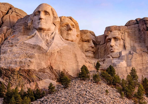 Im warmen Abendlicht ragen die vier Präsidentenköpfe von Mount Rushmore aus den Black Hills in South Dakota über bewaldeten Felsen empor.