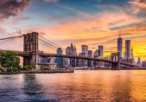 Über dem East River spannt sich die Brooklyn Bridge vor der Skyline von New York im warmen Licht des Sonnenuntergangs mit leuchtenden Wolken. USA-Sehenswürdigkeiten