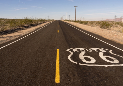 Eine leere Asphaltstraße durch die Wüste zeigt das weiße Route-66-Schild auf der Fahrbahn und führt unter blauem Himmel in die Ferne.