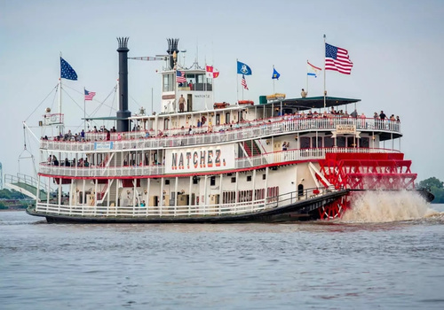 Der Raddampfer Natchez fährt über den Mississippi, während amerikanische Flaggen an Deck wehen und Passagiere die Fahrt verfolgen.