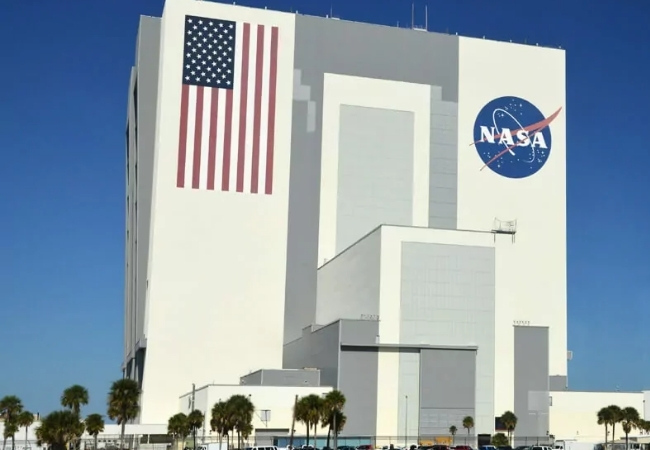 Vor blauem Himmel steht am Kennedy Space Center in Florida ein weißes NASA-Gebäude mit US-Flagge, großem NASA-Logo und Palmen am weitläufigen Gelände.