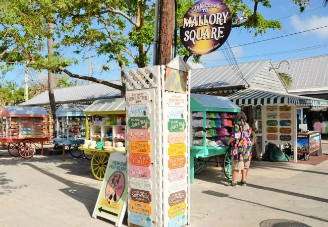 Unter dem Schild von Mallory Square stehen bunte Marktstände in der Sonne, während Besucher zwischen Souvenirs, Hüten und kleinen Verkaufswagen flanieren.