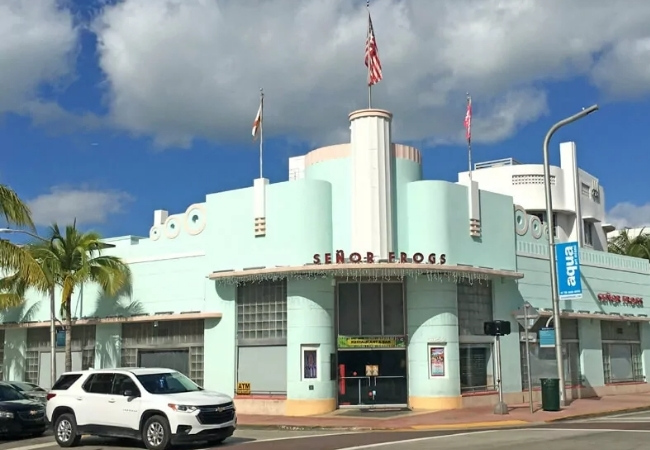 Vor einem pastellfarbenen Art-Deco-Gebäude in Miami steht ein weißer SUV, darüber wehen US-Flaggen unter einem wolkigen Himmel an der Ecke.