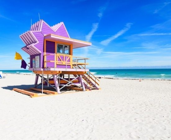 Vor dem türkisblauen Atlantik steht ein pastellvioletter Rettungsschwimmerturm am weißen Sandstrand von Miami Beach unter klarem Himmel.