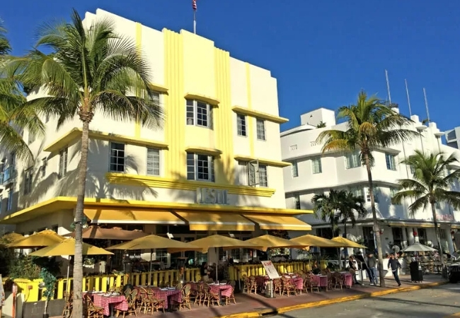 An der Ocean Drive in Miami steht ein gelbes Art-Déco-Gebäude neben Palmen und einem Straßencafé unter klarem blauem Himmel.