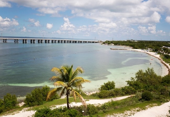 Bahia Honda State Park zeigt eine ruhige Bucht mit hellem Sand, Palmen im Vordergrund und der langen Brücke über dem Wasser.