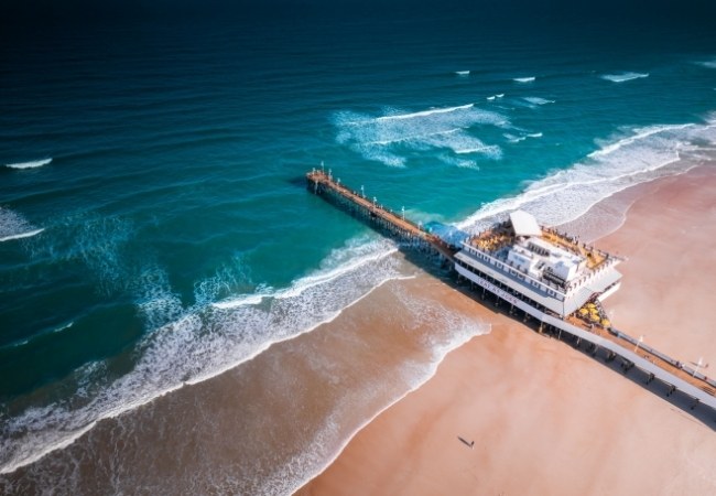 Die lange Seebrücke von Daytona Beach ragt in türkisfarbenes Wasser, während Wellen auf den breiten Sandstrand vor der Küste Floridas treffen.