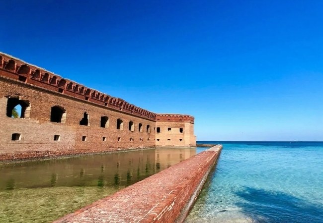 Vor Fort Jefferson im Dry Tortugas National Park treffen rote Backsteinmauern auf klares, türkisblaues Meer unter wolkenlosem Himmel in Florida.