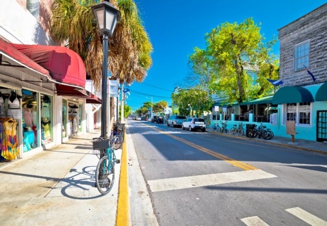 Entlang der Duval Street in Key West stehen bunte Ladenfronten, Palmen und geparkte Fahrräder unter strahlend blauem Himmel.