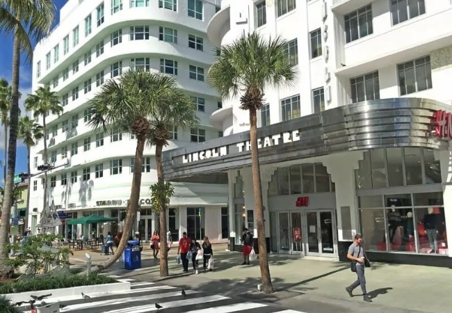 Palmen säumen die Lincoln Road in Miami, vor dem Lincoln Theatre und weißen Art-déco-Gebäuden schlendern Passanten an Geschäften vorbei.