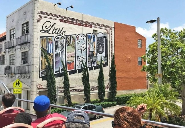 Vor einer bunten Little-Havana-Wandmalerei in Miami fahren Besucher auf einem offenen Bus an Palmen, Autos und einem gelben Fußgängerhinweis vorbei.