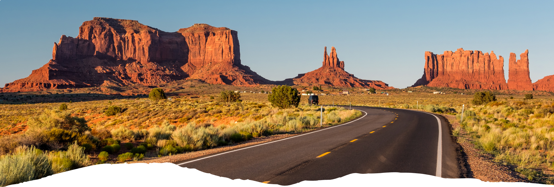 Eine kurvige Asphaltstraße führt durch die rote Wüstenlandschaft mit hohen Tafelbergen im Südwesten der USA bei warmem Abendlicht und weitem Himmel.