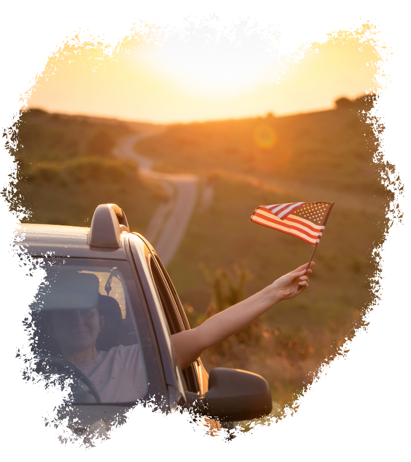 Bei Sonnenuntergang streckt eine Person im Auto eine kleine US-Flagge aus dem Fenster auf einer kurvigen Straße durch hügelige Landschaft.