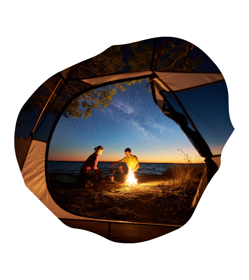 Zwei Reisende sitzen am Lagerfeuer am Seeufer, während der Sternenhimmel über dem Campingplatz in der Abenddämmerung hell leuchtet.