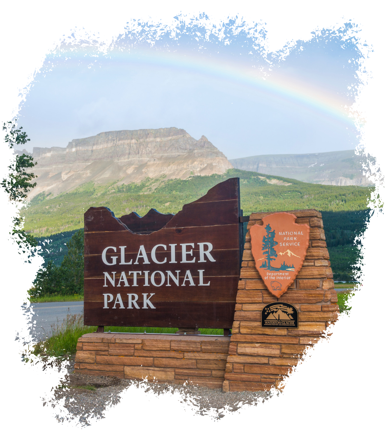 Vor dem Glacier National Park steht ein großes Eingangsschild, dahinter erhebt sich eine grüne Berglandschaft unter einem zarten Regenbogen.