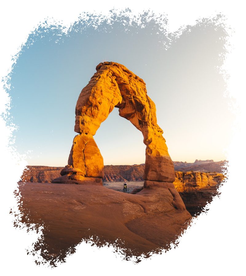 In der warmen Abendsonne leuchtet der Delicate Arch im Arches National Park, während eine einzelne Person klein auf dem Felsplateau darunter steht.