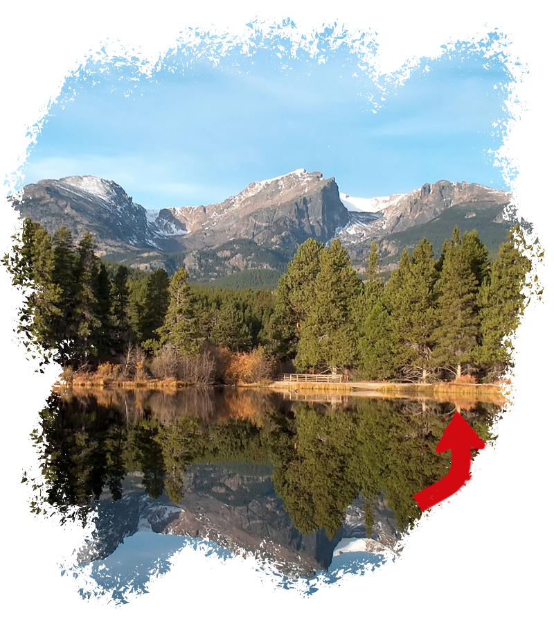 Im Rocky Mountain Nationalpark spiegeln sich schneebedeckte Gipfel und dichte Kiefern im ruhigen See unter blauem Himmel und herbstlichen Uferfarben.