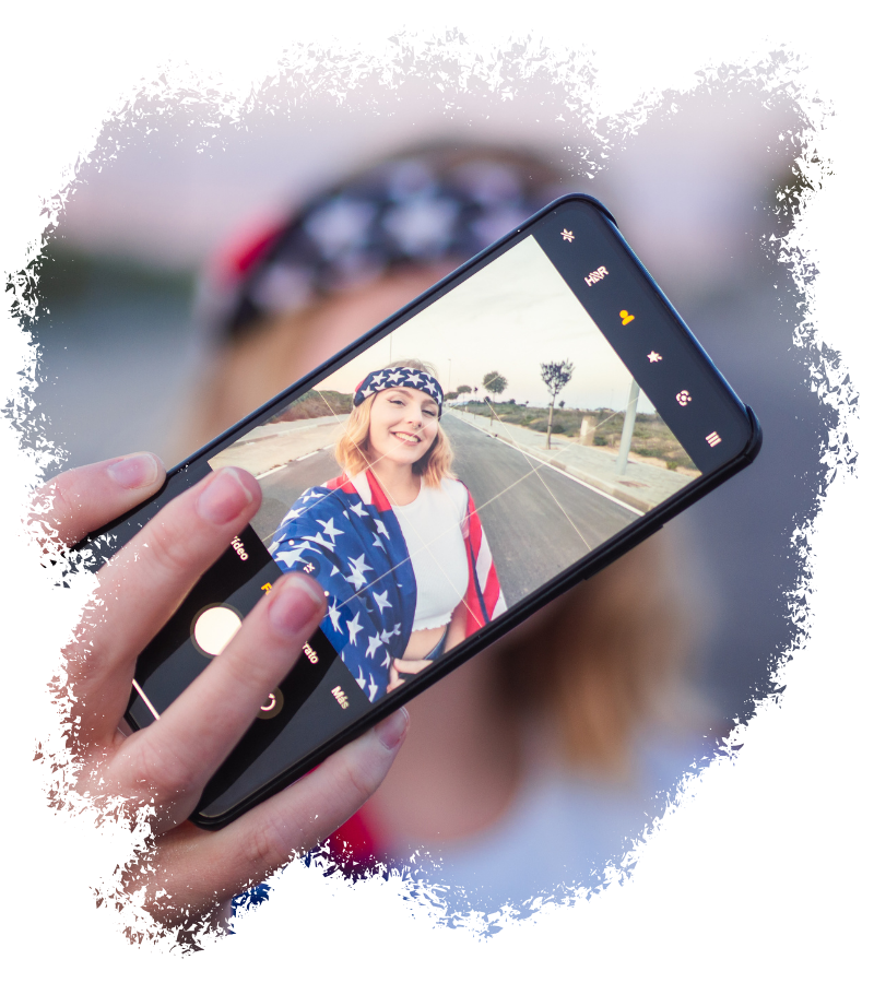Eine junge Frau macht mit dem Smartphone ein Selfie, trägt Stars-and-Stripes-Kleidung und ein Stirnband mit US-Flaggenmuster auf einer Straße.