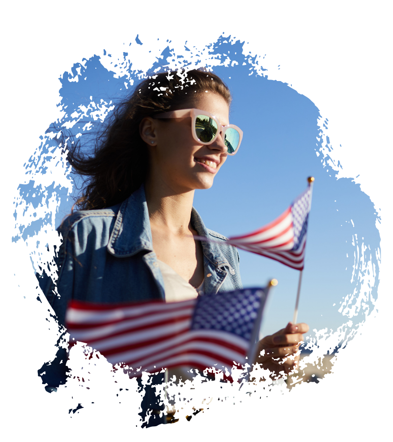Eine lächelnde junge Frau mit Sonnenbrille hält zwei US-Flaggen vor blauem Himmel, während ihr Haar im Wind weht und sommerliche USA-Reise-Stimmung entsteht.