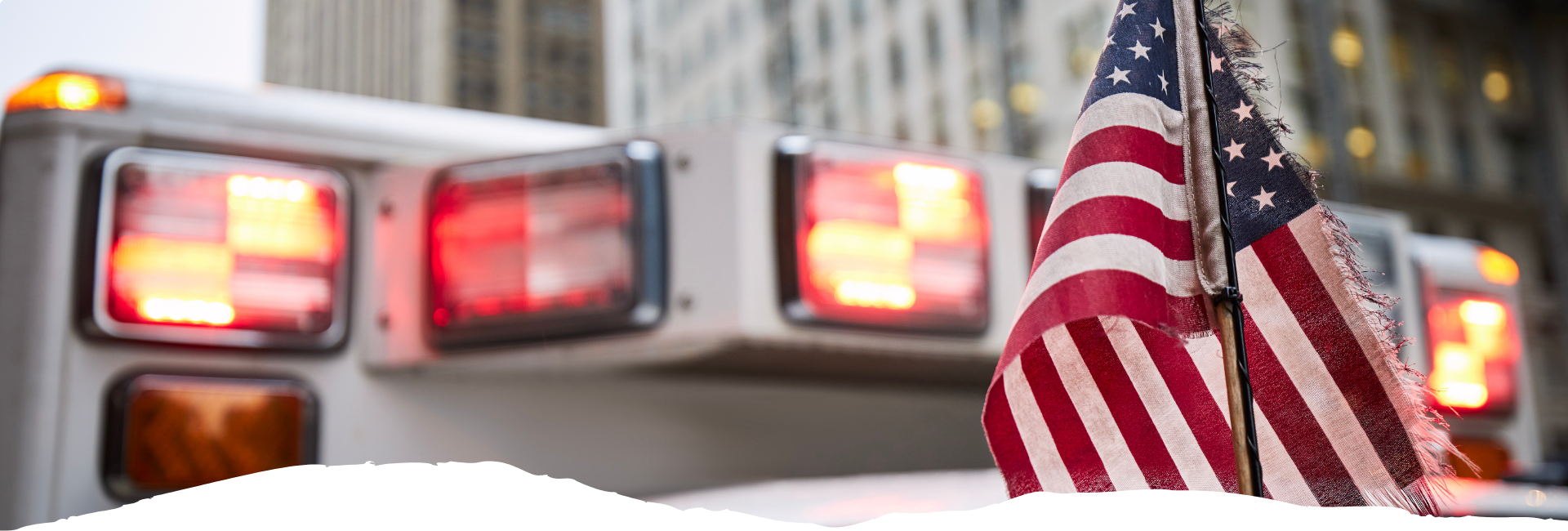 Ein Rettungswagen mit blinkenden Warnlichtern steht in der Stadt, daneben weht eine amerikanische Flagge an einem Mast.