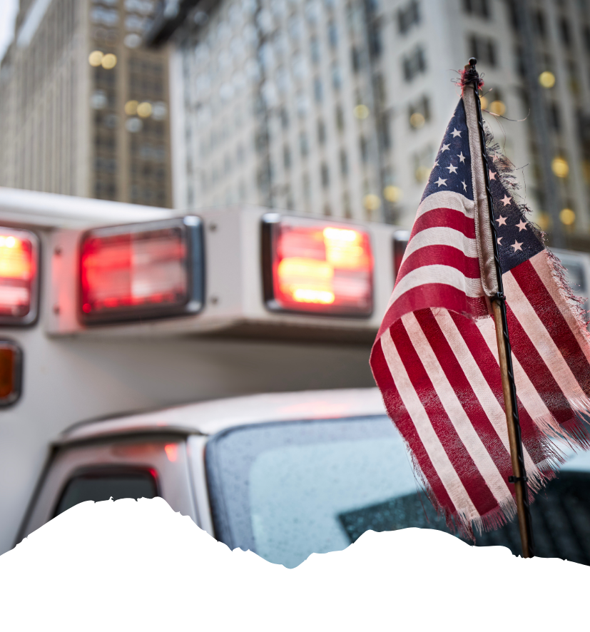 Vor einem Rettungswagen mit blinkenden Warnlichtern weht eine US-Flagge in einer Straßenschlucht zwischen Hochhäusern.