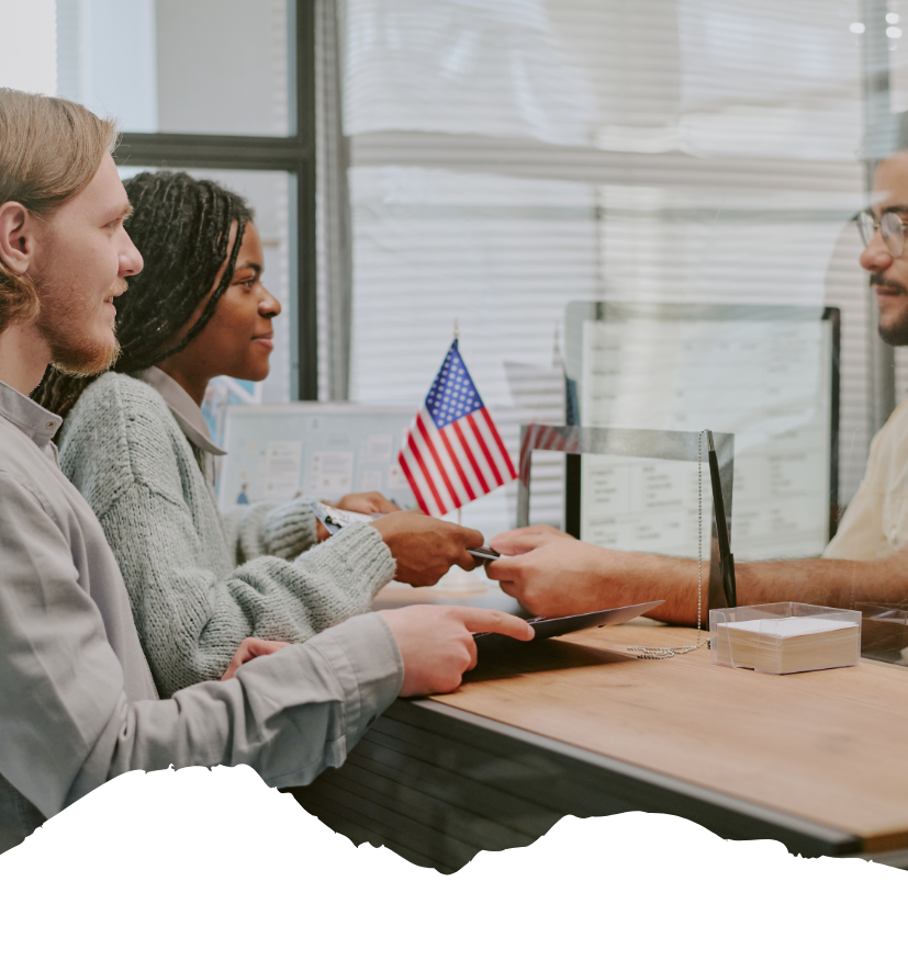 Am Schalter übergeben Reisende ihre Pässe, während ein Beamter hinter der Glaswand die Dokumente prüft und eine kleine US-Flagge im Hintergrund steht.