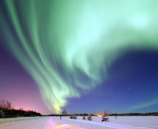 Grüne Polarlichter leuchten über einer verschneiten Straße und einem stillen Winterland unter klarem Nachthimmel in Alaska.