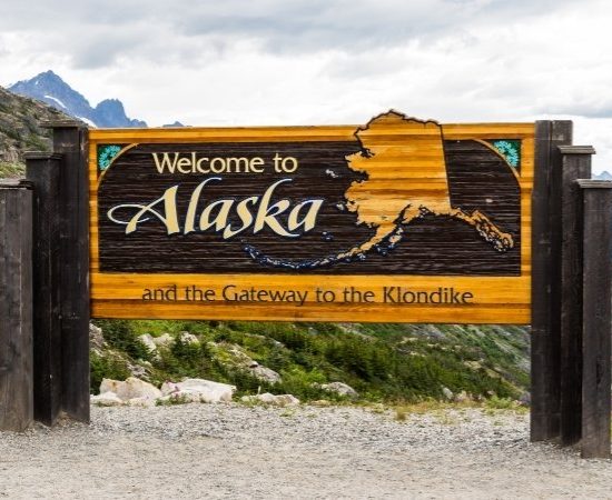 Ein hölzernes Willkommensschild für Alaska steht vor alpiner Berglandschaft und trägt den Hinweis auf den Klondike.