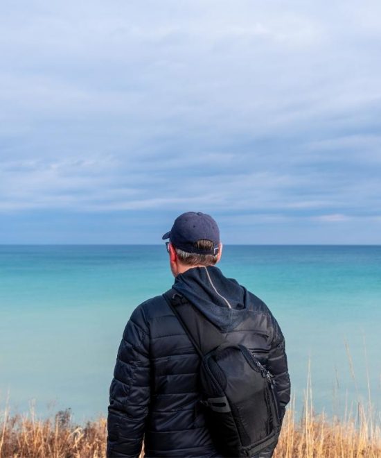 Ein Mann mit Rucksack blickt von einem grasigen Ufer auf den türkisblauen See unter einem wolkigen Himmel. usa reise Great Lakes Rundreise