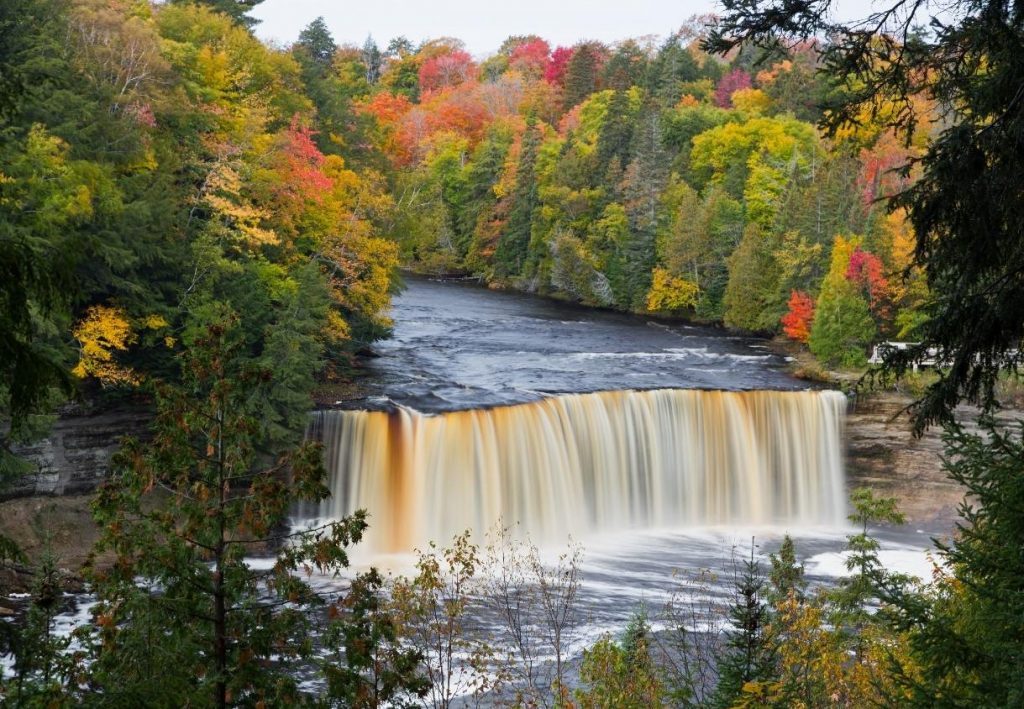 Ein breiter Wasserfall stürzt durch einen herbstlich bunten Wald mit rot, gelb und grün gefärbten Bäumen.