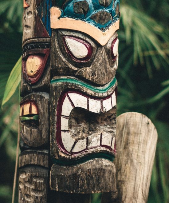Ein bunt bemalter Holztotem mit maskenartigem Gesicht steht vor dichtem tropischen Blattwerk auf Hawaii.