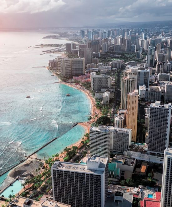 Waikīkīs Skyline in Honolulu reicht bis zur türkisblauen Küste von Oʻahu, während Sonne und Wolken über dem Meer liegen.