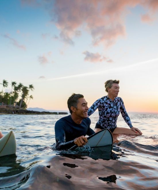 Mehrere Surfer sitzen mit Brettern im Wasser vor Palmen und beobachten den Sonnenuntergang an der Küste von Hawaii.