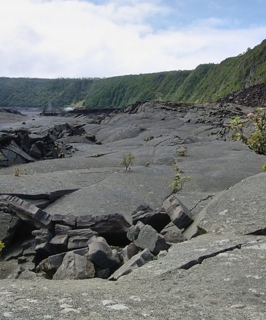 Weite Vulkanlandschaft auf Hawaii mit schwarzer Lava, zerklüftetem Fels und grünen Klippen unter hellem Himmel.