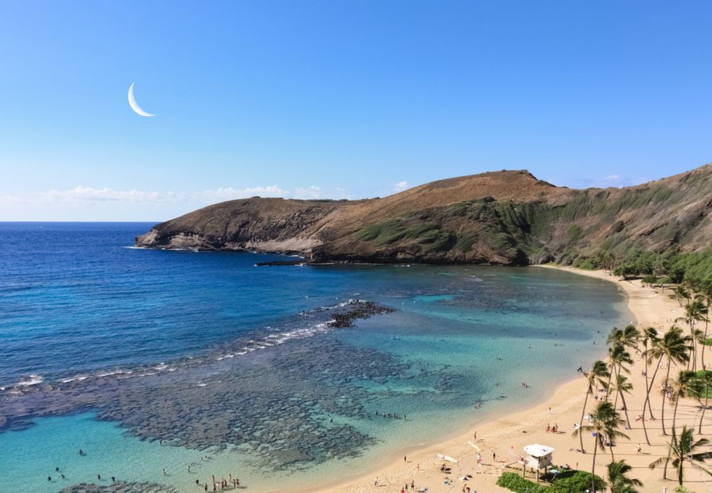 Blick auf eine geschützte Hawaii-Bucht mit türkisblauem Wasser, Sandstrand, Palmen und steilen vulkanischen Hängen.