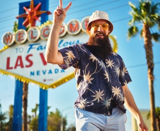 Vor dem Welcome-Schild von Las Vegas steht ein bärtiger Mann mit Hut, Sonnenbrille und Hawaiihemd und zeigt nach oben.
