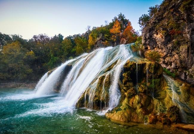 Ein breiter Wasserfall stürzt über moosige Felsen in türkisgrünes Wasser, umgeben von herbstlich verfärbten Bäumen.