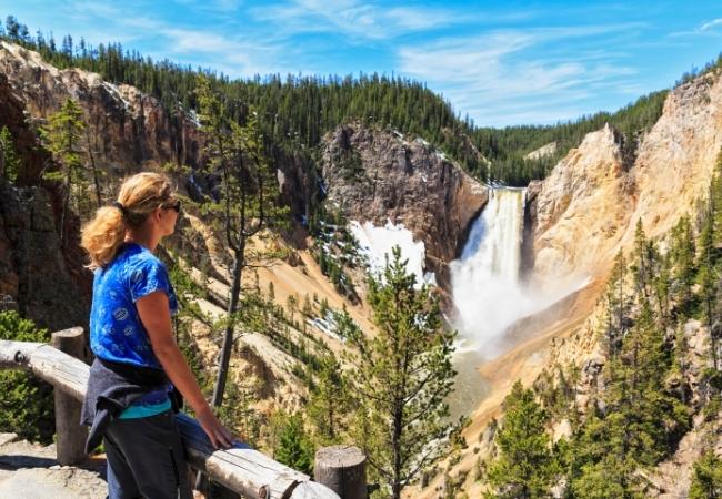 Eine Wanderin blickt über die Schlucht auf einen mächtigen Wasserfall zwischen steilen Felswänden und Kiefern.